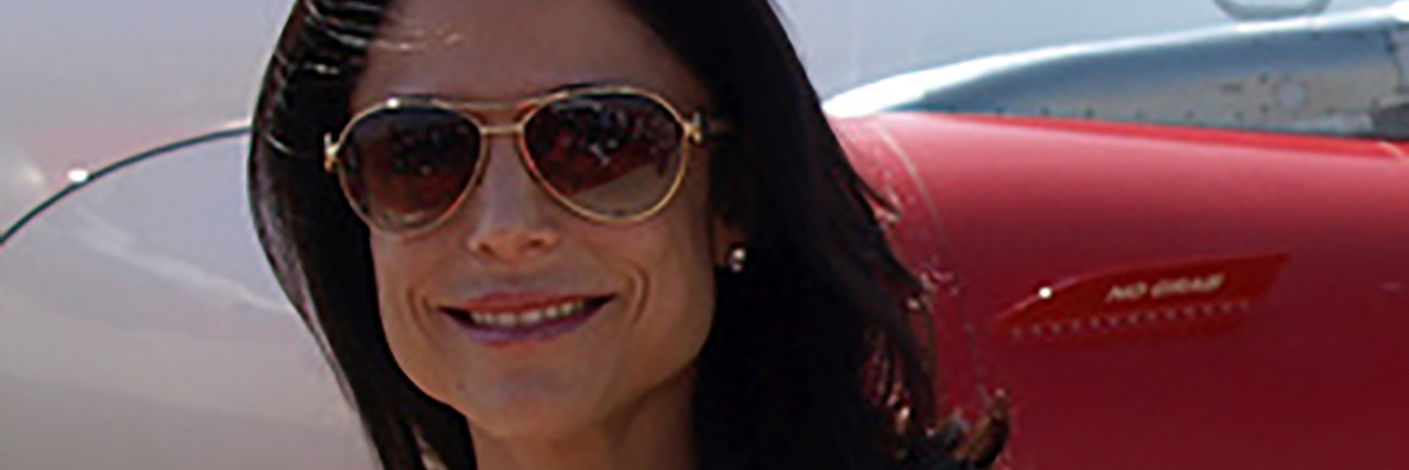 Bethenny Frankel Shamed by Airline Pilot for Fish Allergy Bethanney Franekl smiles while wearing a red dress in front of a plane