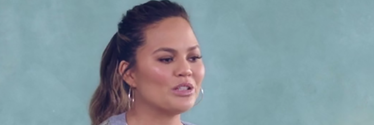 Chrissy Teigen speaks and wears a white dress with a robe