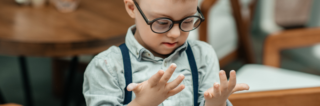 When Your Neurodiverse Child Is Called 'Aggressive' A boy with Down syndrome looks at his hands,. He's wearing glasses and suspenders.
