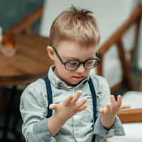 A boy with Down syndrome looks at his hands,. He's wearing glasses and suspenders.