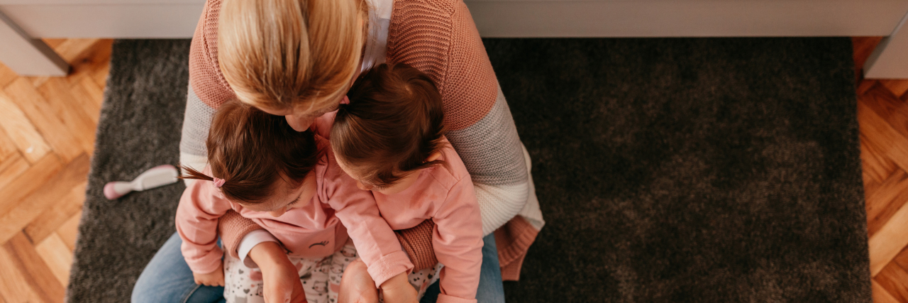 Why I Developed Postpartum Depression After Giving Birth to Twins photo of blonde woman holding twin toddlers on floor with a smartphone