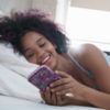 a young black woman in her bed holding her phone and smiling