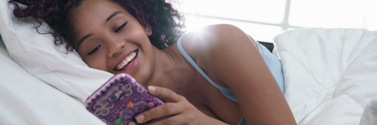 a young black woman in her bed holding her phone and smiling