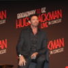 Hugh Jackman in a black suit looks ahead at a press conference