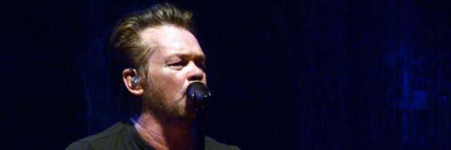 John Mellencamp sings onstage at a concert while playing his guitar