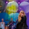 Kesha sings in an all-black outfit in front of a colorful backdrop