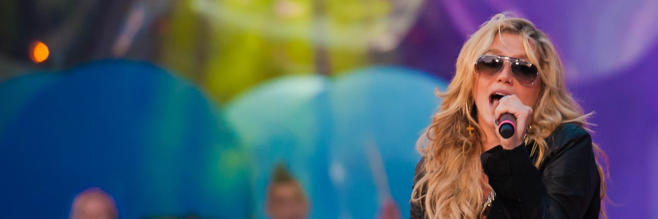 Kesha Writes for Time Magazine About Routines in Mental Health Kesha sings in an all-black outfit in front of a colorful backdrop