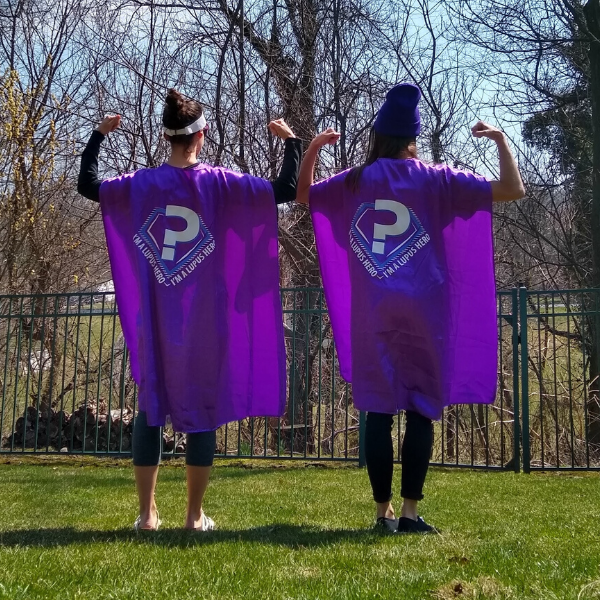 Two women standing outside wearing purple capes. They're standing away from the camera and flexing both arms.