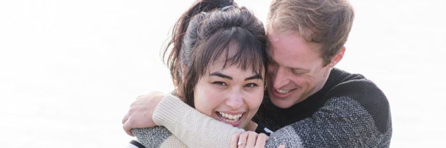 Mickey Rowe and Helen Marion. An Asian woman with bangs and a loose pony tail smiles while in the arms of a man with light brown hair who is looking down.