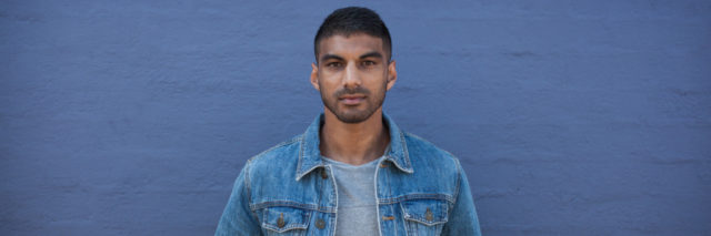 photo from Movember, showing man in denim jacket standing against wall and looking into camera with serious expression