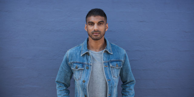 Movember’s Challenge for Checking In With Male Friends During COVID-19 Isolation photo from Movember, showing man in denim jacket standing against wall and looking into camera with serious expression