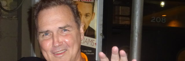 Norm MacDonald poses with a fan outside. He is wearing a bright orange shirt.