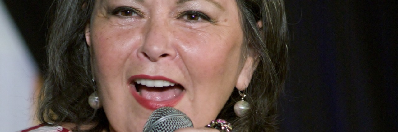 Roseanne Barr Says 'Roseanne' Character Killed By Opioid Overdose on Spinoff 'The Conners' Roseanne Barr in a striped shirt speaks into a microphone