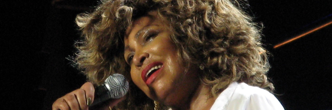 Tina Turner sings onstage while wearing a white ensemble