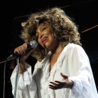 Tina Turner sings onstage while wearing a white ensemble