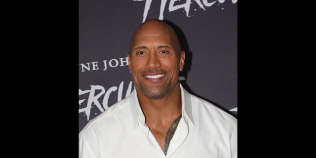 The Rock poses on the red carpet while wearing a white button-down shirt