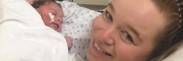 A mother is smiling at the camera while holding her newborn baby in the hospital after birth.
