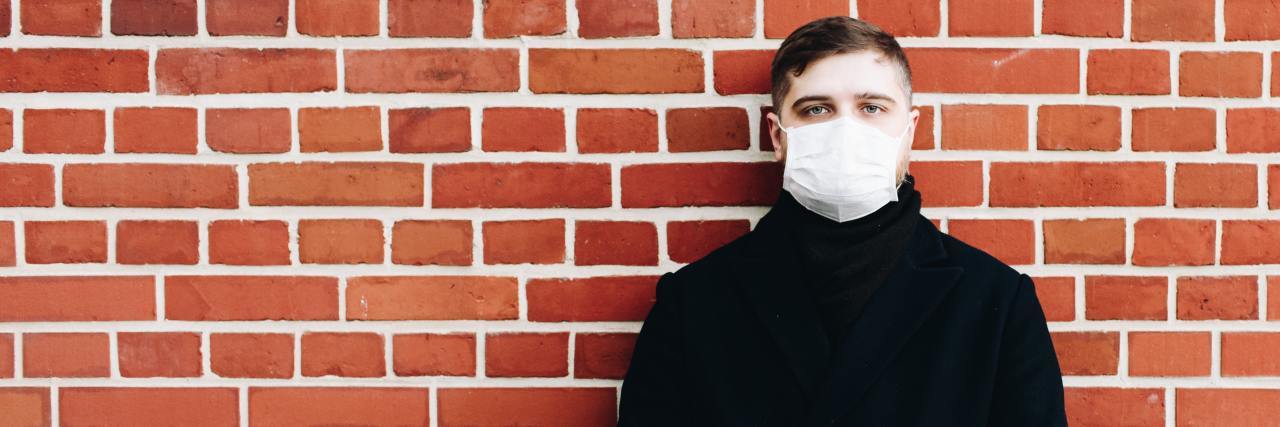 Feeling Lonely and Mental Illness During the Coronavirus Pandemic photo of man in black coat with white face mask, standing in front of brick wall and looking into camera