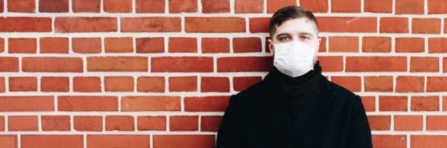 photo of man in black coat with white face mask, standing in front of brick wall and looking into camera