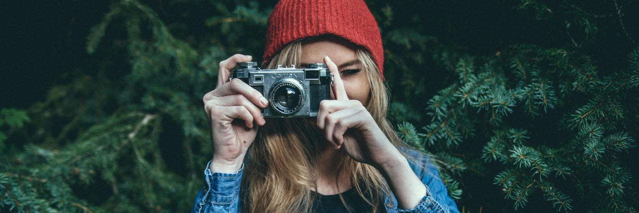 young woman in a red beanie standing in front of a tree taking a picture with a digital camera