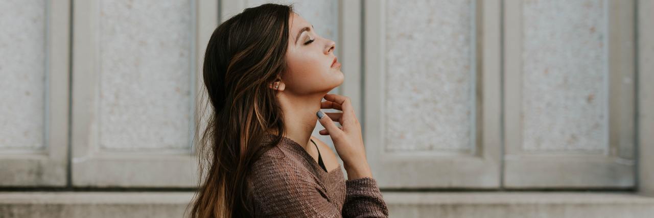 How I've Had to Redefine My 'Normal' During COVID-19 With Anxiety young woman standing outside an off-white building with her head tilted back, eyes closed and hand on her chin