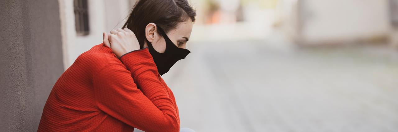 photo of woman in orange knealing in the street against wall while wearing a black face mask
