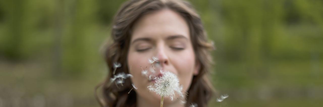 a woman standing outside with her eyes closed, blowing a dandelion