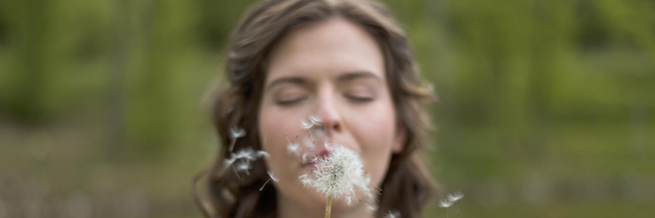 3 Steps You Can Take to Reduce Feelings of Overwhelm a woman standing outside with her eyes closed, blowing a dandelion