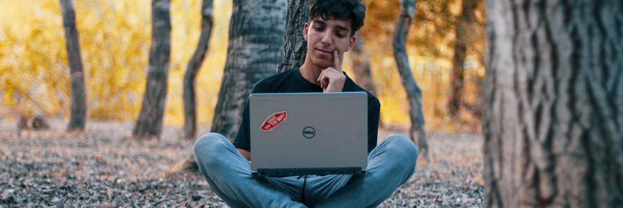 photo of young man in forest, alone, sitting with laptop on his crossed legs and smiling