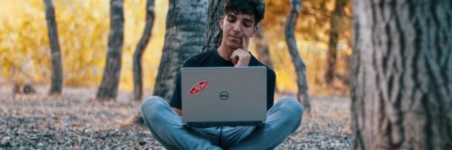 photo of young man in forest, alone, sitting with laptop on his crossed legs and smiling
