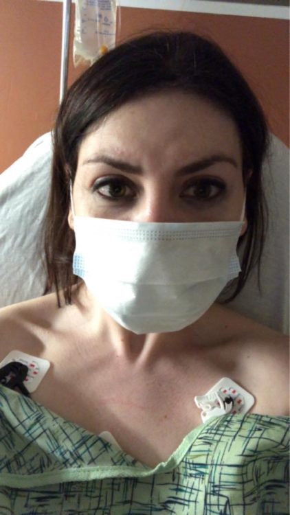 A woman with a face mask and hospital gown on, lying in bed
