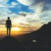 photo of man standing on mountaintop looking at mountains and sunset in distance