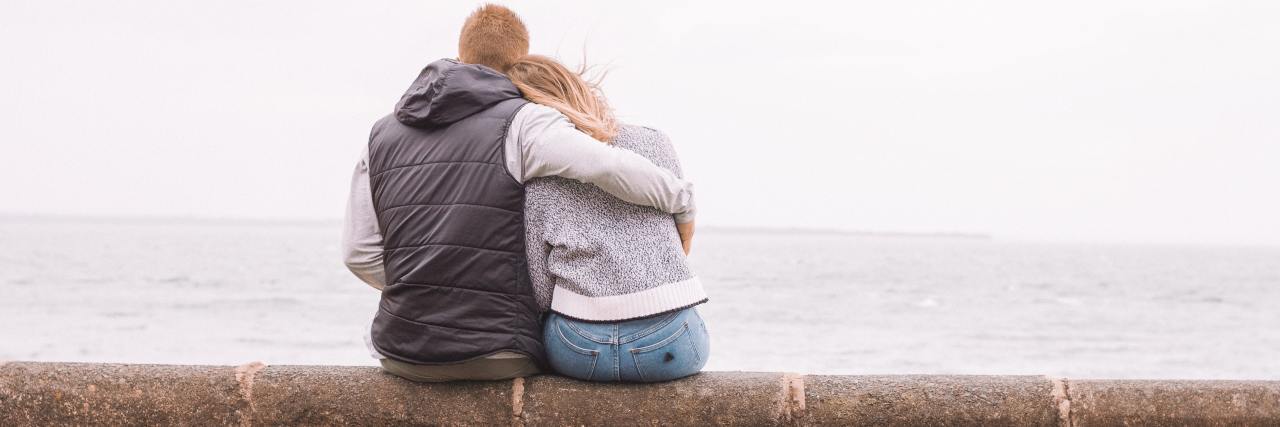 My Partner Helped Me Sit With My Emotions and Prove They're Temporary back of a man with her arm wrapped around his girlfriend's back, both sitting on a log