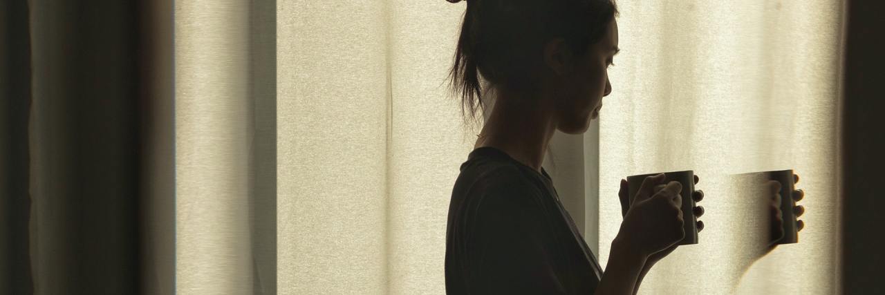 Read This If You Feel COVID-19 Isolation Is Getting to You photo of woman standing by window with a mug