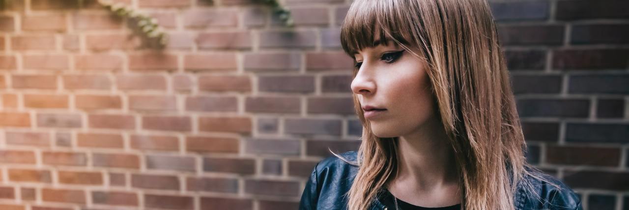 COVID-19 Has Showed Me Being Busy Won't 'Cure' My Mental Illness young adult in a black jacket standing against a brick wall looking down slightly with a straight face