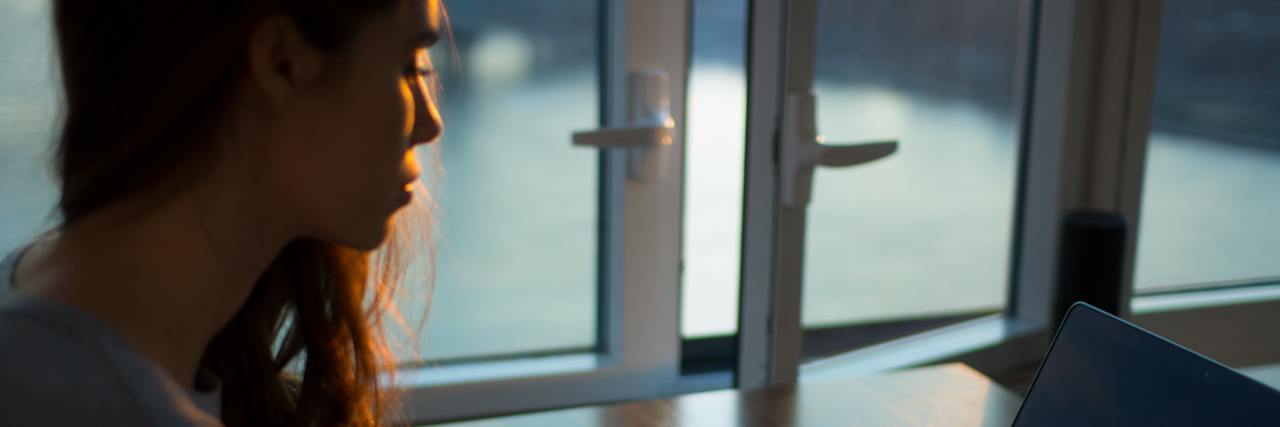Living Through the Coronavirus Pandemic Is Like Trauma photo of woman sitting with laptop by window at sunset