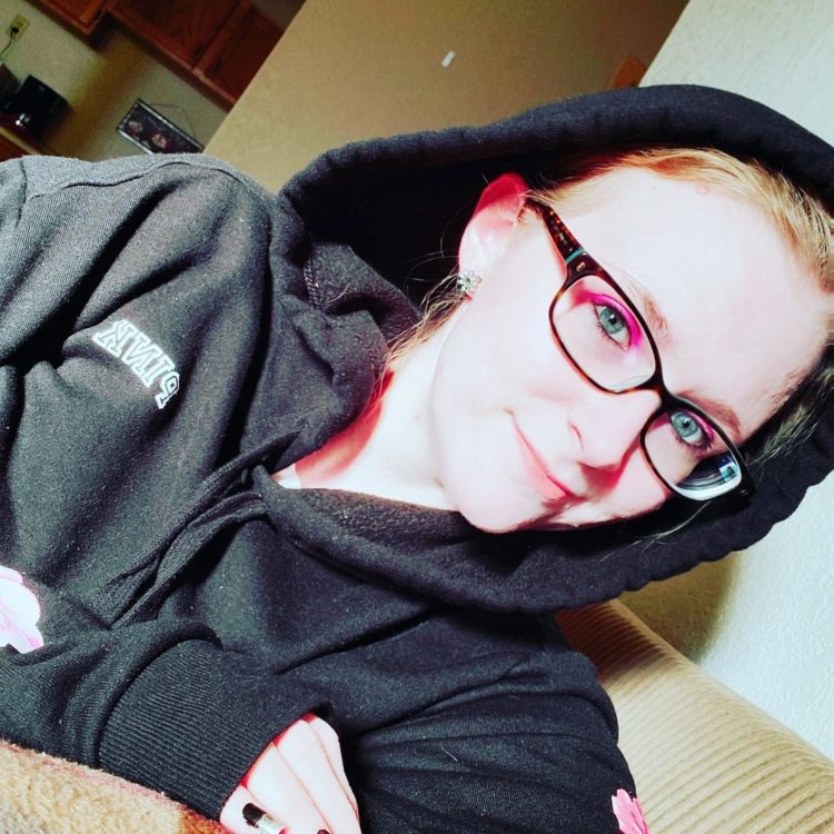A woman with pale skin and blonde hair sits on a couch with a hoodie