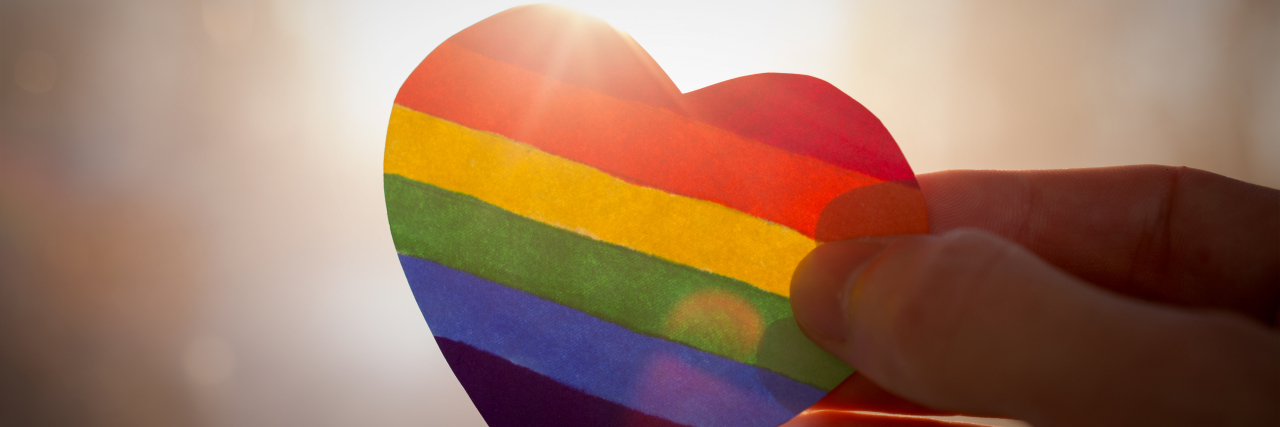 Supporting Your Child Who Just Came Out as Transgender Hand holds a heart painted like a LGBT flag, silhouetted against sun