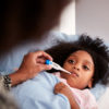 Young black girl lying in bed while a parent holds a thermometer near her mouth