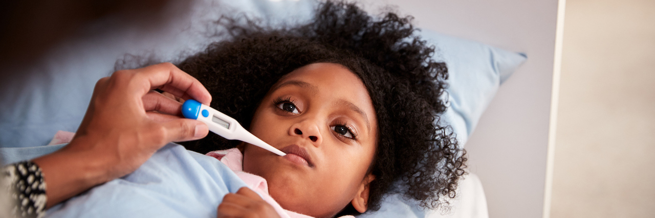 How COVID-19 Symptoms May Present in Kids Young black girl lying in bed while a parent holds a thermometer near her mouth
