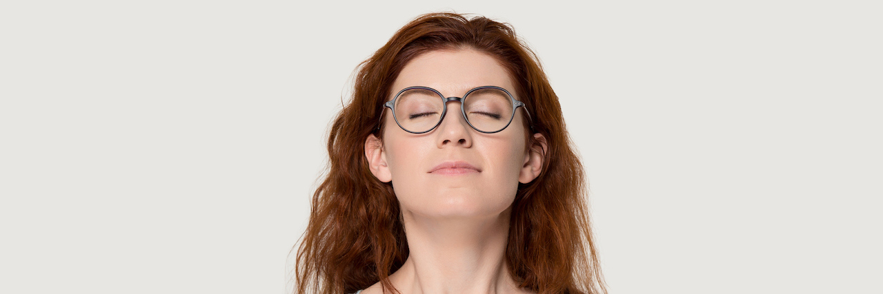 White woman with long red hair, wearing glasses and a light blue T-shirt, has eyes closed, sniffing the air