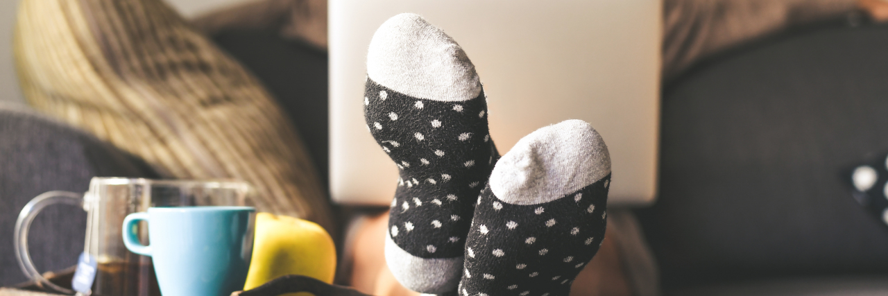 Woman sitting on a couch in the living room with warm socks, coffee and laptop.