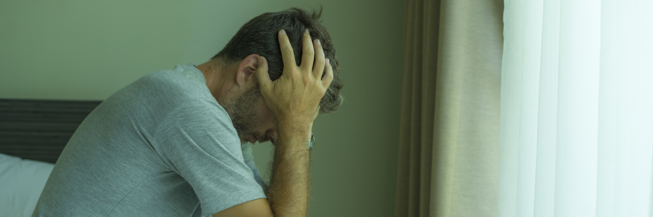 Effects of Loneliness and Being Alone During COVID-19 Pandemic photo of a man with his head in his hands, sitting on the edge of a bed.