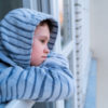 Little boy in gray pajamas looking through window.