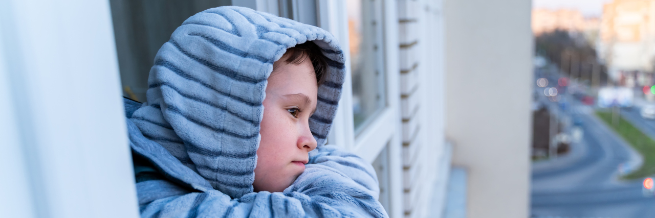 My Child With a Disability Was Isolated Before COVID-19 Little boy in gray pajamas looking through window.