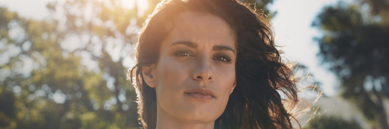 close up of a woman's face, standing in a field with trees, and the sun shining behind her