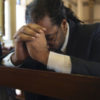 Black man with head bowed in pew at a church with his young daughter standing next to him