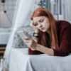 A woman with red hair lying on her bed and looking at her phone with a sad expression on her face