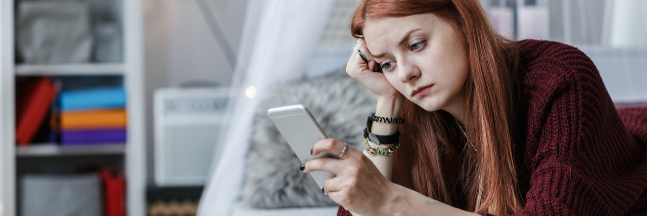 A woman with red hair lying on her bed and looking at her phone with a sad expression on her face