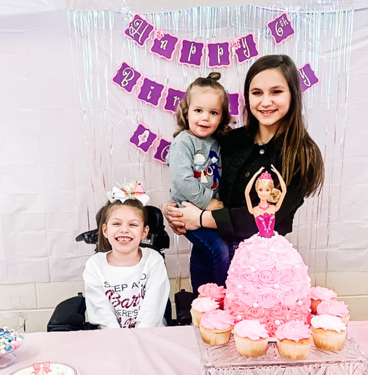 Three young sisters are sitting near a birthday cake at a party.